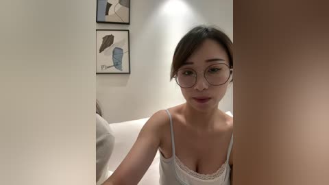 Video of an Asian woman with fair skin, glasses, and light brown hair, wearing a white lace camisole. She leans against a beige wall adorned with abstract art.