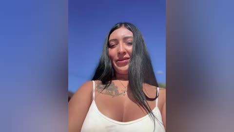 Video of a young woman with long black hair, medium skin tone, and a tattoo on her chest, wearing a white tank top, against a clear blue sky background.