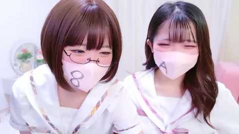 Video of two young Asian women in white masks, glasses, and floral-patterned dresses, indoors with a pink couch and a vase with flowers in the background.