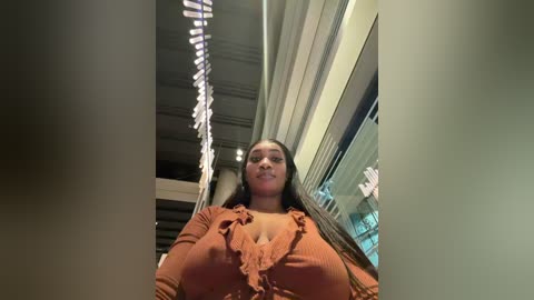 Video of a young Black woman with long braids, wearing a low-cut, ruffled, brown top, standing in a modern, well-lit mall corridor with glass storefronts and metal railings.