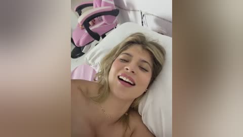 Video of a topless, fair-skinned blonde woman with a joyful expression, lying on a white pillow, surrounded by pink and white furniture in a bedroom.