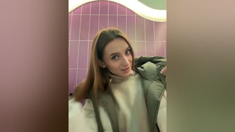 Media: Video of a young Caucasian woman with straight, light brown hair, wearing a beige sweater and a green jacket, standing in front of a pink tiled wall with a curved design.
