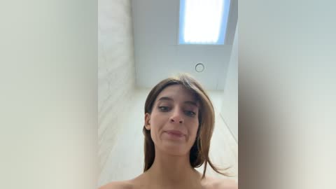 Video of a young, fair-skinned woman with straight brown hair, standing nude in a white-walled room with a small, bright window.