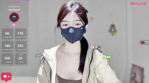 A video of an East Asian woman with light skin, straight black hair, and wearing a black mask, beige jacket, and glasses, in a modern, minimalist room.