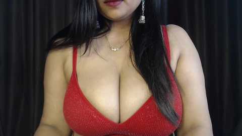 Video of a woman with long black hair, wearing a low-cut, red, sequined dress, adorned with dangling earrings and a delicate necklace. She stands against a dark, textured curtain background.