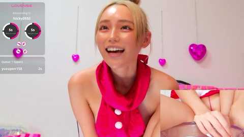 Video of a smiling Asian woman with blonde hair in a ponytail, wearing a bright pink sleeveless top with buttons, in a pink room with heart decorations.