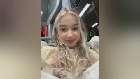 Video of a young woman with long, wavy, platinum blonde hair, wearing a light yellow sweater, sitting in a modern bus. The background includes a gray interior with yellow wires and a red emergency exit sign.