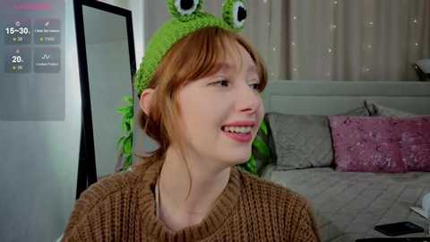 Video of a young woman with fair skin and red hair, smiling, wearing a green frog hat and a brown knitted sweater, sitting in a cozy bedroom with white bedding and a pink pillow.