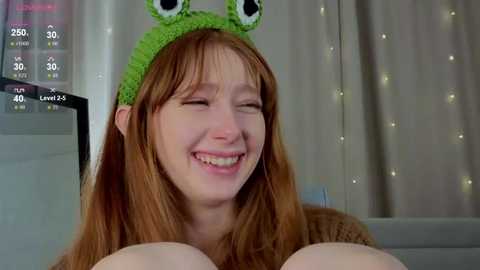Video of a smiling, fair-skinned woman with long red hair wearing a green frog hat, holding large, bare breasts, in a cozy room with white curtains.