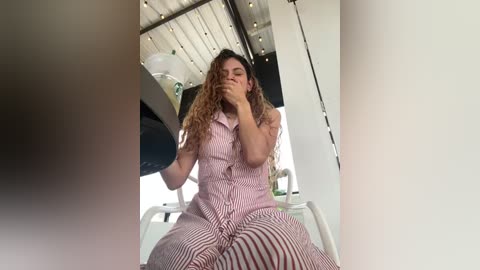 Video of a woman in a pink and white striped dress, sitting on a white chair, covering her mouth with her hand, in a modern, well-lit room with white walls and a white ceiling.