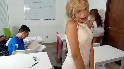 Video of a classroom with a blonde woman in a white dress standing in front, a man in a blue shirt seated, and another woman in the background.