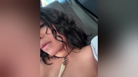 Video of a young woman with medium skin tone, short curly black hair, and a light-colored necklace, seen from the side, in a car.