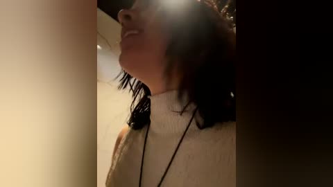 Video of a smiling woman with short dark hair, wearing a white turtleneck, standing indoors against a beige wall, with a blurry background.