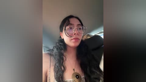 Video of a young woman with long black hair, wearing glasses, a fur collar, and a large pendant, sitting in a car.