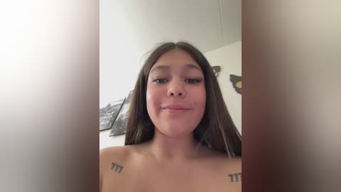 A video of a young woman with long brown hair, fair skin, and a slight smile, standing indoors. She has tattoos on her shoulders and is topless.