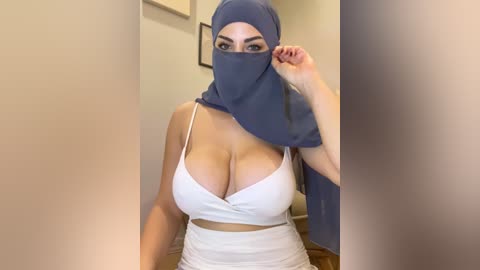 Video of a woman with large breasts wearing a white crop top, blue hijab, and high-waisted white pants, in a hallway with beige walls and framed art.