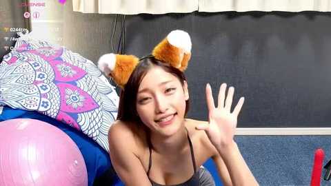 A smiling East Asian woman with short black hair, wearing a gray top and fox ear headband, lies on a blue couch with a pink and white mandala cushion.