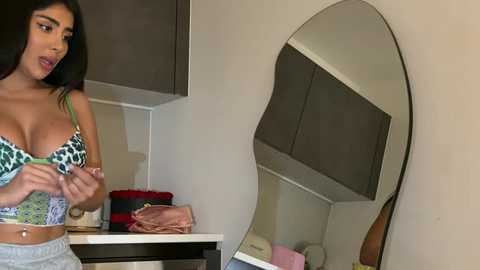 Video of a Latina woman with long black hair, wearing a leopard-print bra and grey sweatpants, adjusting her bra in a modern kitchen with dark cabinets and a round mirror.