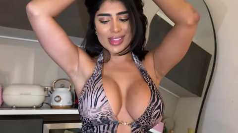 Video of a curvy, dark-haired woman with large breasts, wearing a leopard print halter top, posing in a modern kitchen with white cabinets and appliances.