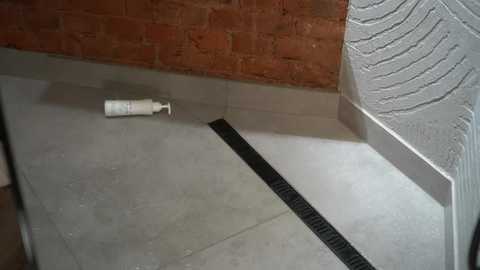 Video of a minimalist bathroom with grey tiles, a black drain, white soap dispenser, and exposed brick wall.