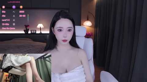 Video of an Asian woman with long black hair, wearing a white towel, sitting on a white chair in a modern bedroom with dark curtains, a TV showing Chinese text, and a bed.