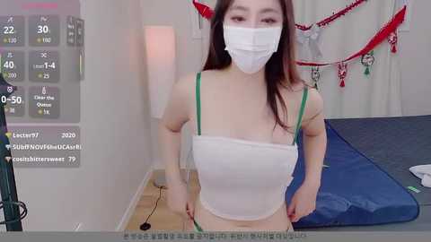 A video of an East Asian woman with long dark hair, wearing a white mask, white tube top, and green bra, standing in a bedroom with a blue bed, red Christmas decorations, and a clock displaying 20:00.