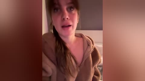 A video of a young woman with light skin, brown hair, and a medium build, wearing a beige hoodie, looking surprised or shocked, partially obscured by a blurred figure in the foreground.