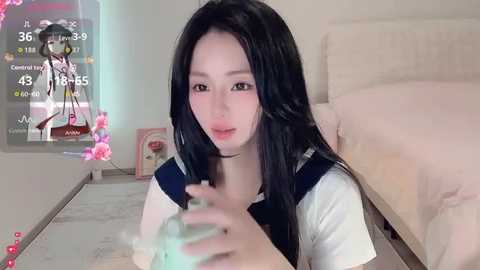 A video of a young Asian woman with long black hair, wearing a white and navy school uniform, holding a green plush toy, sitting on a beige carpet in a modern bedroom with a bed, framed photo, and a digital clock displaying 3:39.