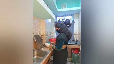 Video of a woman in a small, brightly lit kitchen with teal walls and patterned tiled backsplash, preparing food. She wears a dark green apron over a long-sleeved shirt, and has long, dark hair.