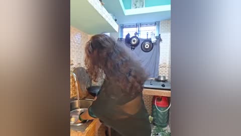 Video of a woman with long, curly hair, wearing a black shirt, washing dishes in a kitchen with a tiled backsplash, hanging pots, and a green laundry basket.