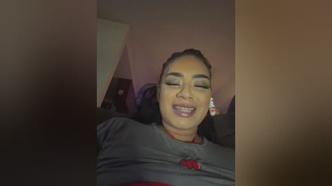 Video of a young woman with medium brown skin and dark hair, smiling widely with braces, wearing a gray shirt. She is indoors with dim lighting, and the background is blurred.