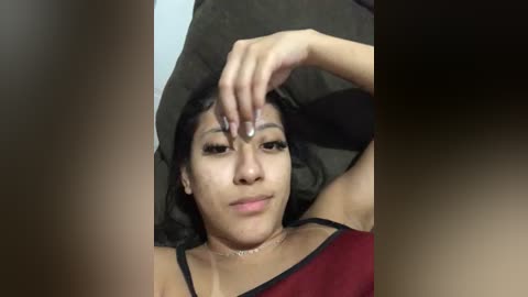 Video of a young woman with light brown skin, long black hair, and a red top, lying on a dark surface, touching her forehead, appearing relaxed.