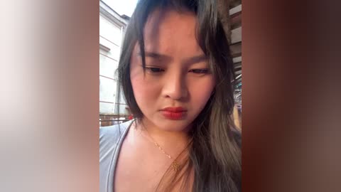 Video of an Asian woman with long black hair, wearing a light blue top and red lipstick, standing in an urban setting with wooden structures in the background.