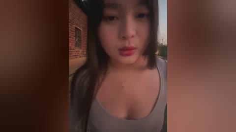 A video of a young Asian woman with straight black hair, wearing a grey tank top, standing in front of a brick wall. She has fair skin and full lips.