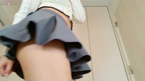 Video of a young woman's lower body, wearing a short, pleated gray skirt and white knit sweater, partially lifting her skirt to reveal a glimpse of her bare thigh. The background shows a beige wall and a closed door.