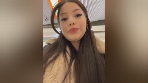 A video of a young Asian woman with long, straight black hair and fair skin, wearing a beige coat, indoors with a blurred background.