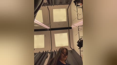 Video of a woman with long, straight hair, wearing glasses, standing in a narrow, dimly lit room with four small, rectangular windows and metal railings.