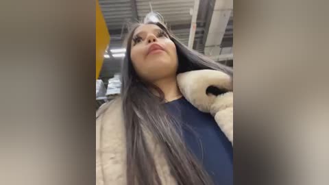 Media: Video of a young woman with long, straight, dark hair, wearing a blue top and a beige fur coat, standing in a warehouse with exposed beams and yellow accents.