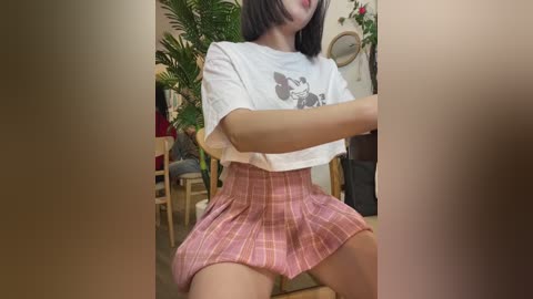 A video of a young woman with medium skin tone and straight black hair, wearing a cropped white shirt and a high-waisted pink plaid skirt, sitting on a chair in a cozy indoor setting with plants and wooden furniture.