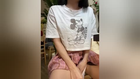 Video of an Asian woman with black hair, wearing a cropped white Mickey Mouse t-shirt and plaid skirt, sitting indoors with potted plants in the background.