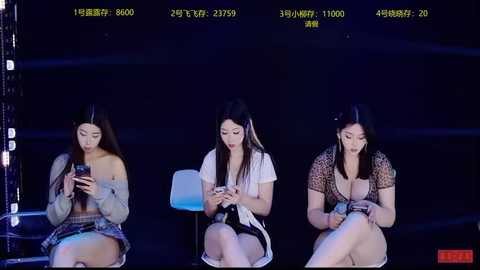 Video of three Asian women with long hair, dressed in casual outfits, seated on a dark stage, all focused on smartphones, illuminated by stage lights.