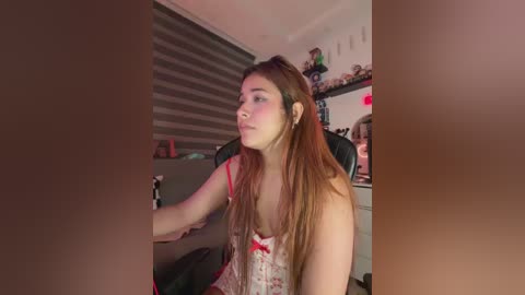 Video of a young woman with long brown hair, wearing a floral camisole, sitting on a black office chair in a dimly lit room with grey vertical blinds and gaming equipment in the background.