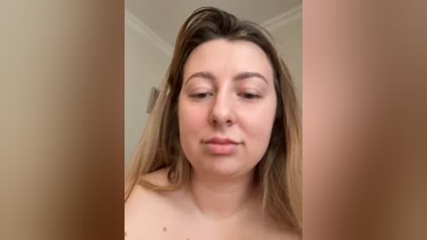Video of a fair-skinned woman with long, straight brown hair, wearing no clothing, in a beige room with a white ceiling.