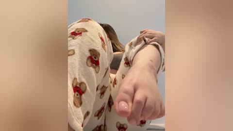 Video of a person with fair skin, wearing light beige pajama pants with teddy bear prints, lying on their side, with a foot extended towards the camera.