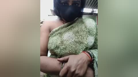 Media: Video of a South Asian woman in a green sari with white floral patterns, black mask, and bracelets, taken indoors with a blurred background.