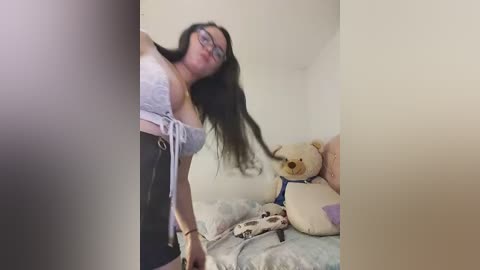 Video of a woman with long, dark hair, wearing glasses, a gray bra, and high-waisted pants, leaning over a bed with a stuffed animal, in a dimly lit, white-walled room.
