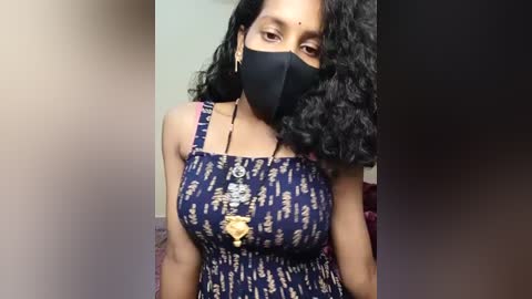 Video of a woman with medium brown skin, wearing a black face mask, black tank top with a gold pendant, and curly black hair, indoors with blurred background.
