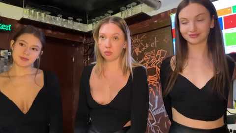 Media: Video of three young women with fair skin and long hair, wearing black tops, in a dimly lit bar with colorful wall art and glassware.