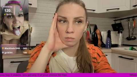 Video of a young woman with long blonde hair, wearing a white t-shirt under an orange-patterned jacket, sitting in a modern kitchen. In the background, a live stream video shows a woman sticking out her tongue.