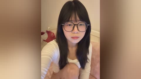 A video of an Asian woman with straight black hair, wearing black-framed glasses, a white top, and red lipstick, sitting on a bed with a red teddy bear.